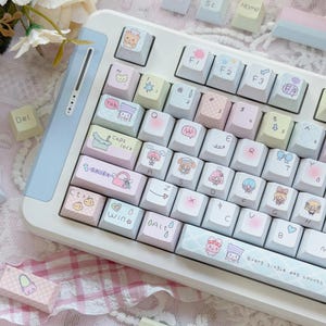 May include: A white and light blue keyboard with custom keycaps featuring pastel illustrations. The keycaps have various designs, including characters, patterns, and text. The keyboard has the text "Every single day counts" on the spacebar.