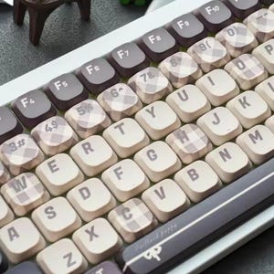 Puede incluir: Primer plano de un teclado mecánico con teclas blancas y grises. Las teclas presentan una mezcla de colores lisos y un patrón de cuadros. Las teclas de función son de color gris oscuro. La barra espaciadora es gris oscuro con el texto "Baillard puppy".