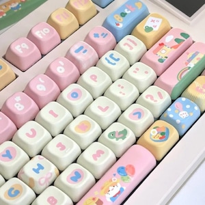 May include: A pastel-colored keyboard with custom keycaps. The keycaps feature a variety of designs, including rainbows, animals, and other cute illustrations. The keyboard has a small screen and a light pink frame. The keycaps are in shades of pink, blue, yellow, and white.