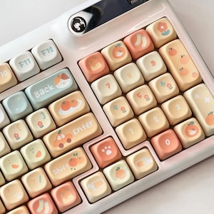 Może przedstawiać: Biała klawiatura z niestandardowymi klawiszami w pastelowych odcieniach. Klawisze ozdobione są ilustracjami pomarańczy, brzoskwiń i jasnoniebieskich elementów. Klawiatura posiada port USB i pokrętło głośności.