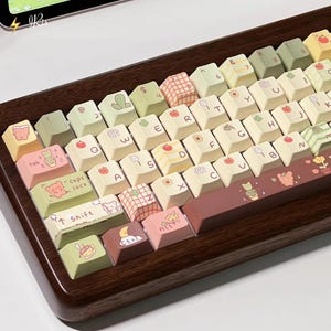 May include: A wooden-framed keyboard with custom keycaps. The keycaps are a mix of cream, green, and pink, decorated with fruit, floral, and animal illustrations. The space bar and shift keys have unique designs. The keyboard has a modern, aesthetic design.