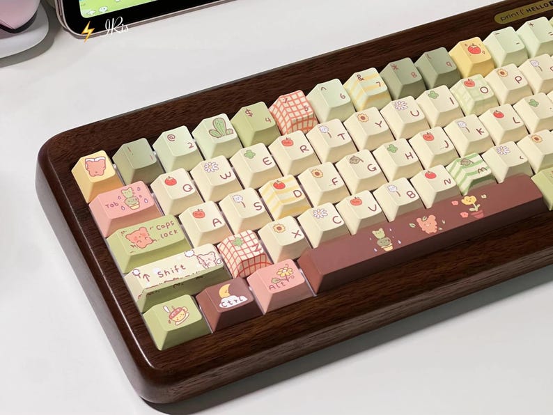 May include: A wooden-framed keyboard with custom keycaps. The keycaps are a mix of cream, green, and pink, decorated with fruit, floral, and animal illustrations. The space bar and shift keys have unique designs. The keyboard has a modern, aesthetic design.