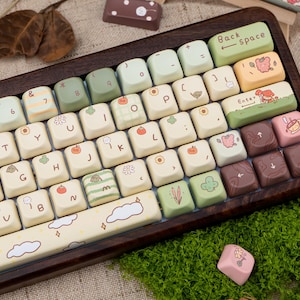 May include: A custom keyboard with a wooden frame and cream-coloured keycaps. The keycaps feature various illustrations, including fruits, clouds, and floral designs. The keyboard has a Backspace and Enter key, and the overall aesthetic is whimsical and decorative.