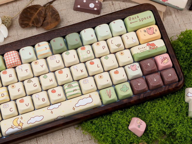 May include: A custom keyboard with a wooden frame and cream-coloured keycaps. The keycaps feature various illustrations, including fruits, clouds, and floral designs. The keyboard has a Backspace and Enter key, and the overall aesthetic is whimsical and decorative.