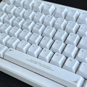 Może przedstawiać: Zbliżenie białej klawiatury mechanicznej z białymi nakładkami na klawisze. Klawisze są ułożone w standardowym układzie QWERTY. Niekt&oacute;re klawisze mają małe dekoracyjne obrazy i tekst. Klawiatura ma białą ramkę i dekoracyjny wz&oacute;r na spacji.
