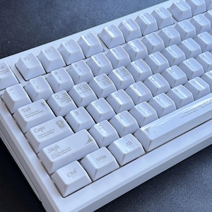 Może przedstawiać: Biała klawiatura mechaniczna z białymi nakładkami na klawisze. Klawiatura ma standardowy układ QWERTY z klawiszami funkcyjnymi i klawiaturą numeryczną. Klawisze są czyste, a klawiatura jest w dobrym stanie.