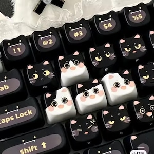 May include: A keyboard with custom keycaps featuring cat faces. The keycaps are black and white with various expressions, including surprised, angry, and happy. Some keys have numbers and symbols, while others have letters.