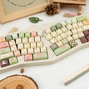 May include: A split ergonomic keyboard with custom keycaps. The keycaps are cream-coloured with pastel green, pink, and brown accents. Some keys feature illustrations of animals and patterns. A matching wrist rest is also present.