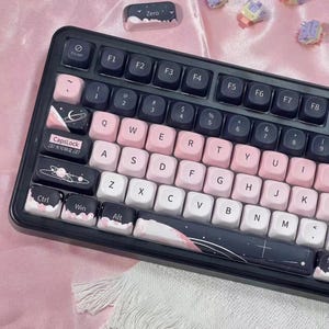 May include: A black and pink mechanical keyboard with white and pink keycaps. The keycaps have a space theme with planets and stars. The keyboard has a black frame and a variety of function keys.