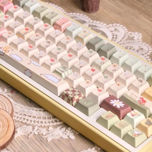 May include: A custom mechanical keyboard with pastel-coloured keycaps. The keycaps are decorated with fruit, cloud, and floral designs. The keyboard has a yellow frame and a white base, creating a soft, aesthetic appearance.