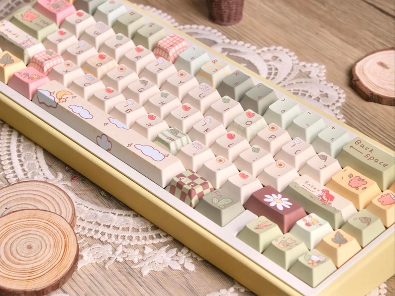 May include: A custom mechanical keyboard with pastel-coloured keycaps. The keycaps are decorated with fruit, cloud, and floral designs. The keyboard has a yellow frame and a white base, creating a soft, aesthetic appearance.
