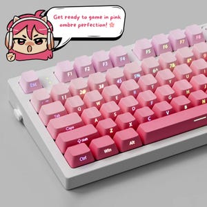 K&ouml;nnte beinhalten: Eine wei&szlig;e mechanische Tastatur mit rosa Ombre-Tastenkappen. Die Tastenkappen gehen von hellrosa in einen tieferen Farbton &uuml;ber, mit wei&szlig;er Beschriftung. Die Tastatur hat eine Cartoon-Grafik und den Text "Get ready to game in pink ombre perfection!"