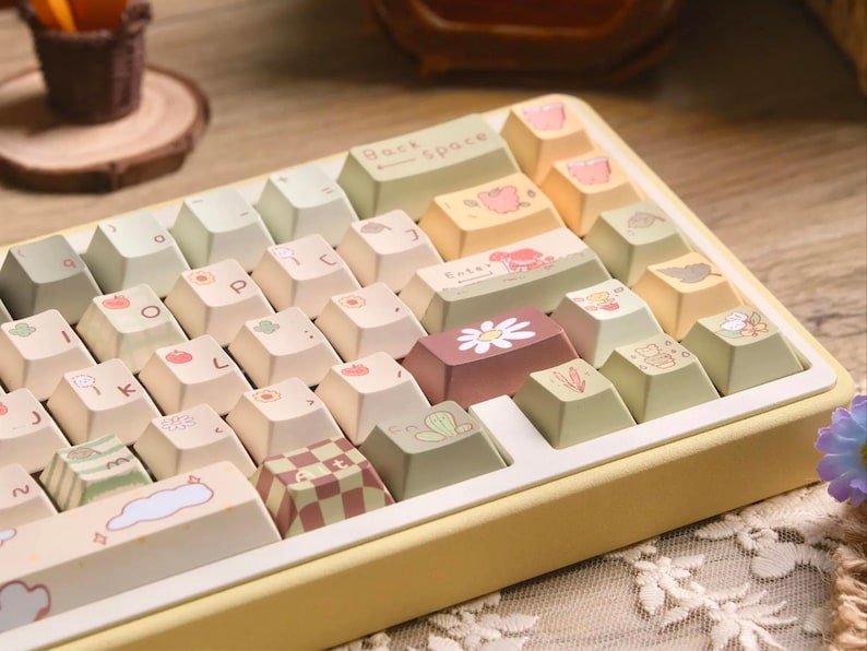 May include: A pastel-coloured mechanical keyboard with custom keycaps. The keycaps feature various designs, including a daisy, a cloud, and a checkered pattern. The keyboard has a yellow frame and a Backspace key.