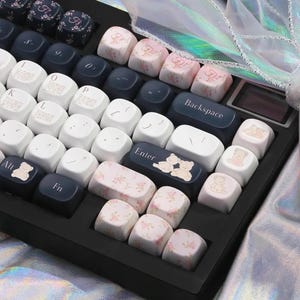 May include: A keyboard with custom keycaps. The keycaps feature a variety of designs, including floral patterns, teddy bear illustrations, and letters. The color scheme includes white, navy blue, and pink.