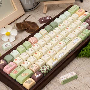 May include: A custom keyboard with a wooden frame and colourful keycaps. The keycaps feature various designs, including cartoon characters, patterns, and text. The keyboard includes a long space bar with a cloud and moon design. The keycaps are in shades of green, pink, and brown.