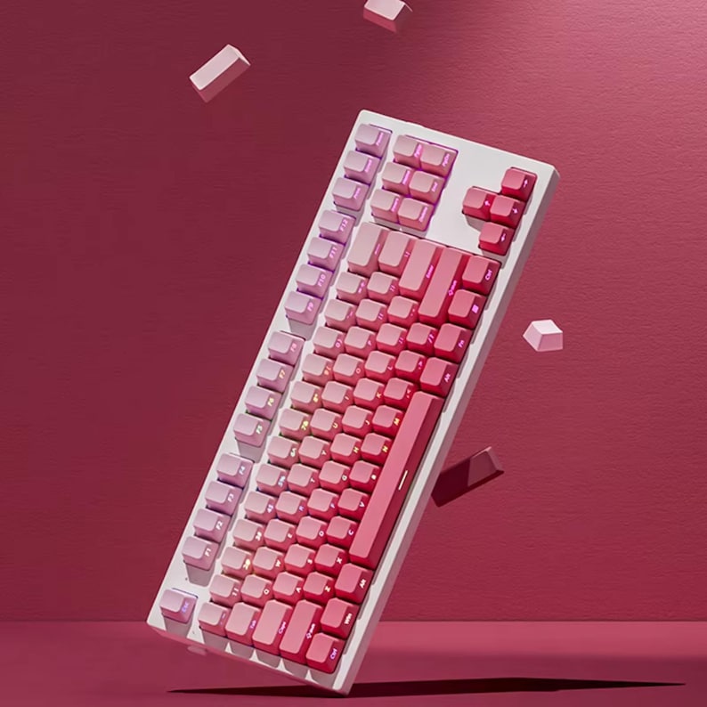 K&ouml;nnte beinhalten: Eine wei&szlig;e mechanische Tastatur mit rosa und roten Tasten. Die Tasten gehen von hellrosa zu tiefrot &uuml;ber und erzeugen einen Farbverlauf. Mehrere lose Tastenkappen sind vor einem roten Hintergrund verstreut.