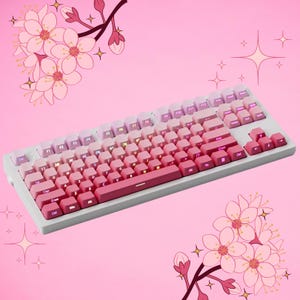 K&ouml;nnte beinhalten: Eine wei&szlig;e mechanische Tastatur mit rosa und lila Tastenkappen in einem Farbverlauf. Die Tastatur hat einen hellgrauen Rahmen und befindet sich vor einem rosa Hintergrund mit Kirschbl&uuml;ten-Illustrationen.