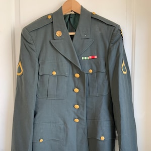 May include: A vintage olive green military jacket with gold buttons and insignia. The jacket features multiple pockets, a notched collar, and a ribbon with red, white, and green stripes. The jacket is hanging on a wooden hanger.