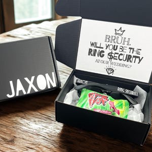 May include: Black gift box with the name "JAXON" in white lettering. The open box contains sunglasses, a watermelon-flavored candy, and a card that reads "BRUH. WILL YOU BE THE RING SECURITY AT OUR WEDDING?"