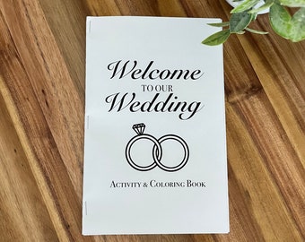 Könnte beinhalten: Ein weißes Heftchen mit dem Text "Welcome to our Wedding" und einer Schwarzweißillustration von zwei ineinander verschlungenen Ringen mit einem Diamanten an einem Ring. Der Text "Activity & Coloring Book" ist unter der Illustration gedruckt.