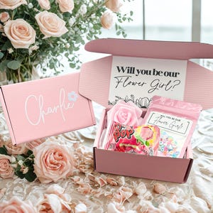 May include: Pink gift boxes with floral and candy contents. One box reads "Charlie" with a flower graphic. The other box is open, revealing a rose, candy, and a card that says "Will you be our Flower Girl?"