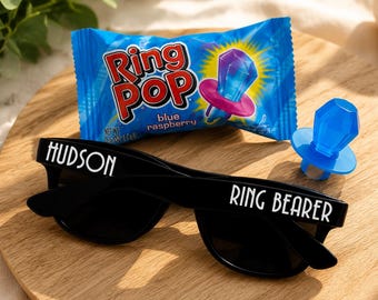 May include: Black sunglasses with the text "HUDSON" on one arm and "RING BEARER" on the other. A blue Ring Pop candy and a blue raspberry Ring Pop candy package are also on a wooden surface.