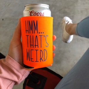 May include: An orange can cooler with the text "Hmm... That's Weird" in blue. The cooler is holding a silver Coors beer can. The image is taken from a slightly elevated angle.