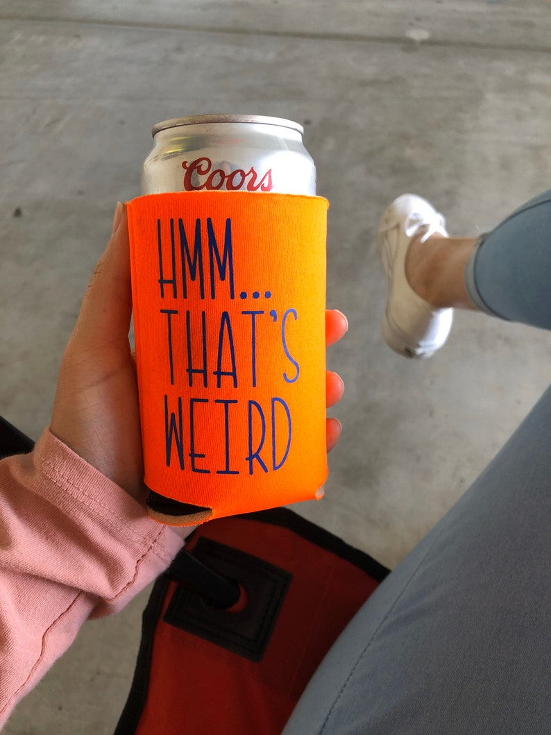 May include: An orange can cooler with the text "Hmm... That's Weird" in blue. The cooler is holding a silver Coors beer can. The image is taken from a slightly elevated angle.