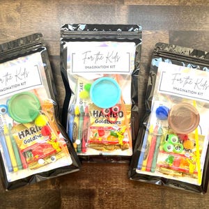 May include: Three clear plastic bags filled with children's craft supplies and snacks. Each bag has a label that reads "For the Kids Imagination Kit." Contents include crayons, candy, and small toys. The bags are sealed with a black zipper.