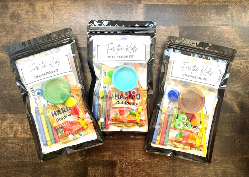 May include: Three clear plastic bags filled with children's craft supplies and snacks. Each bag has a label that reads "For the Kids Imagination Kit." Contents include crayons, candy, and small toys. The bags are sealed with a black zipper.