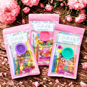 May include: Three pink zipper bags labeled "For the Kids IMAGINATION KIT" containing colorful craft supplies and Haribo Goldbears candy. The bags are arranged on a wooden surface with pink flowers in the background.