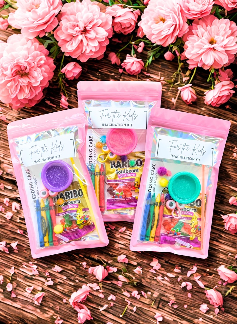 May include: Three pink zipper bags labeled "For the Kids IMAGINATION KIT" containing colorful craft supplies and Haribo Goldbears candy. The bags are arranged on a wooden surface with pink flowers in the background.