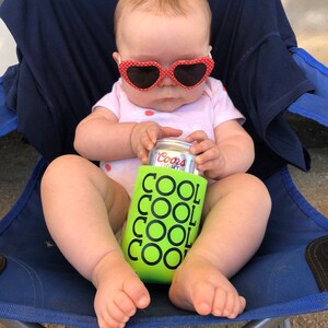 May include: A baby wearing heart-shaped sunglasses and a pink onesie sits in a blue chair. The baby holds a can of Coors Light in a green can cooler with the words "COOL" repeated. The image is taken outdoors.