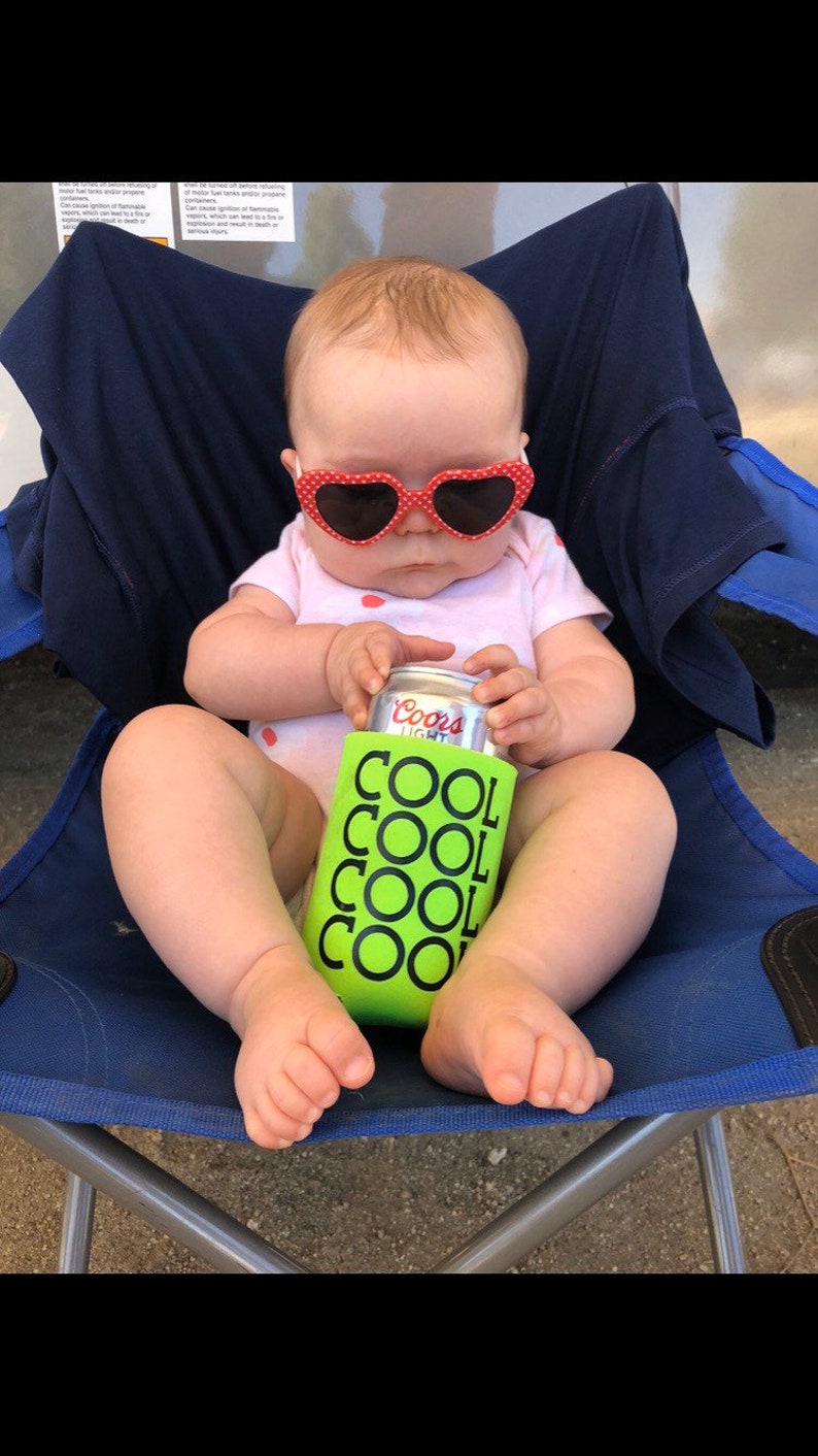 May include: A baby wearing heart-shaped sunglasses and a pink onesie sits in a blue chair. The baby holds a can of Coors Light in a green can cooler with the words "COOL" repeated. The image is taken outdoors.