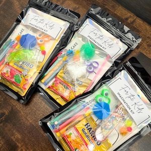 May include: Three clear plastic bags labeled "For the Kids IMAGINATION KIT" containing various craft supplies and candy. Contents include colorful pens, pom-poms, googly eyes, and Haribo Goldbears. The bags are sealed and arranged on a wooden surface.