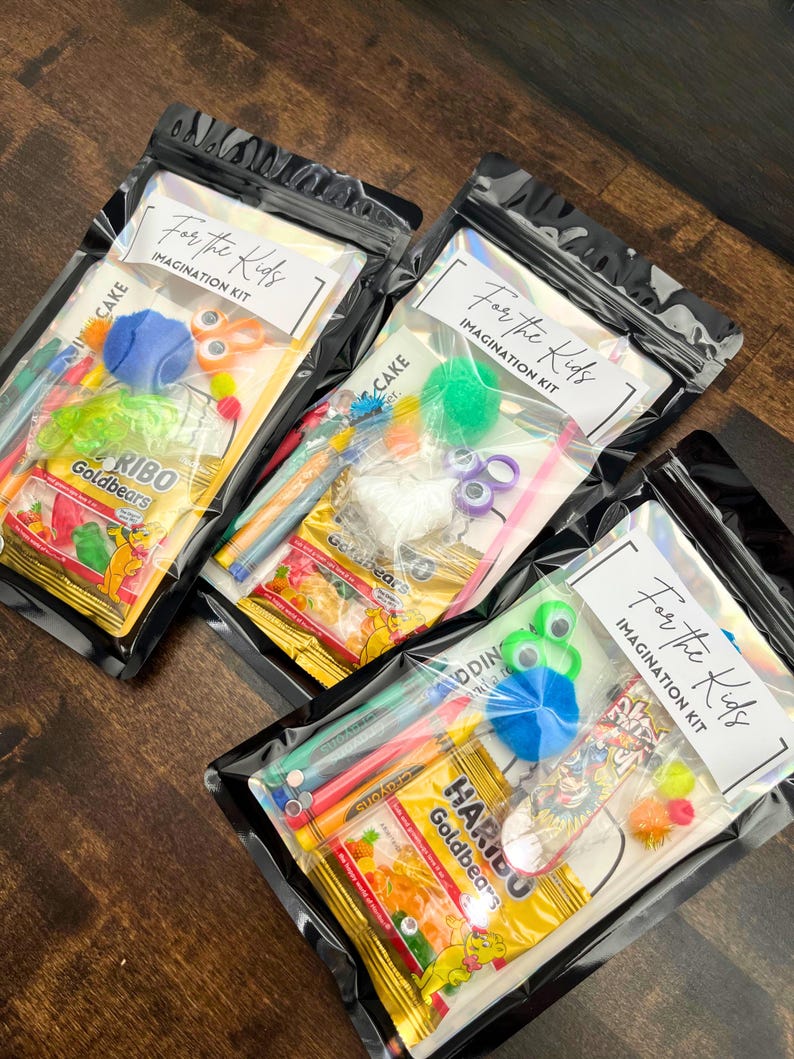 May include: Three clear plastic bags labeled "For the Kids IMAGINATION KIT" containing various craft supplies and candy. Contents include colorful pens, pom-poms, googly eyes, and Haribo Goldbears. The bags are sealed and arranged on a wooden surface.