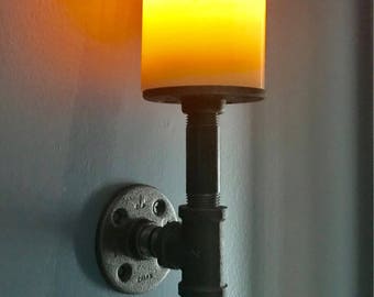 Könnte beinhalten: Eine schwarze Wandleuchte aus Metallrohren mit einem runden, orange beleuchteten Kerzenhalter. Die Leuchte ist an einer Wand mit hellblauem Hintergrund montiert.