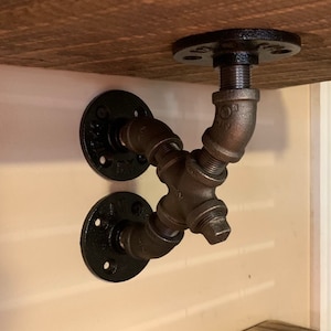 May include: Two black metal pipe shelf brackets with flanges. The brackets are connected with a T-shaped pipe fitting.