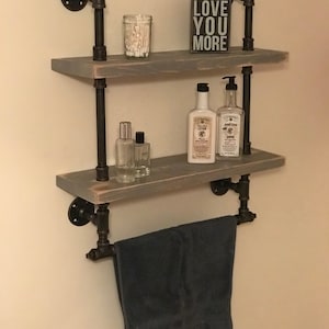May include: Two-tiered wooden shelf with black metal pipe supports. The shelf is decorated with bottles of lotion, a jar of cotton swabs, and a sign that reads "Love You More". A black towel is hanging on a pipe towel bar.