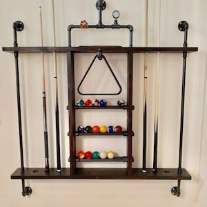 May include: A wall-mounted pool cue rack made of dark wood and black metal pipes. The rack has shelves for storing pool balls and a triangle rack for holding the balls in a pyramid shape. There are four cues hanging on the rack.