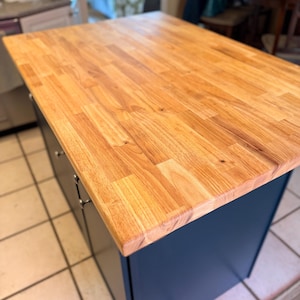 Puede incluir: Una isla de cocina con encimera de bloque de carnicero y base azul marino. La encimera está hecha de tablas de madera de color claro. La isla tiene cajones y puertas de armario con herrajes plateados. La isla está sobre un suelo embaldosado.