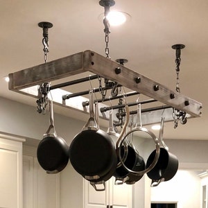 May include: A rustic wooden pot rack with black metal pipes and chains hanging from the ceiling. The rack is holding a variety of pots and pans.