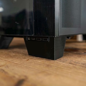 May include: A black computer tower case featuring a front-facing panel with a headphone jack and two USB ports. The chassis has a matte black finish with a perforated side vent and is supported by sturdy, angled black feet at the base.