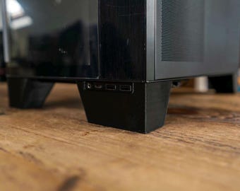 May include: A black computer tower case featuring a front-facing panel with a headphone jack and two USB ports. The chassis has a matte black finish with a perforated side vent and is supported by sturdy, angled black feet at the base.