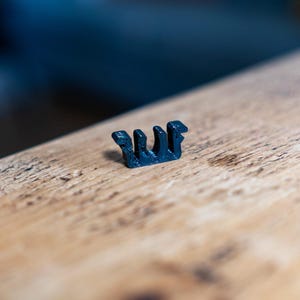 May include: A small, dark blue plastic clip with a unique, stylized design. The clip is positioned on a light brown wooden surface, with a blurred blue background. The clip's design features multiple vertical elements.
