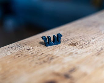 May include: A small, dark blue plastic clip with a unique, stylized design. The clip is positioned on a light brown wooden surface, with a blurred blue background. The clip's design features multiple vertical elements.