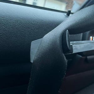 May include: A close-up shot of a black car seat belt. The belt is made of a woven material and is secured by a black plastic buckle. The surrounding interior features black leather-like material.