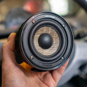Peut inclure: Un haut-parleur audio de voiture noir avec un centre imitation bois et un dôme central noir. Le haut-parleur porte les inscriptions "MADE IN FRANCE" et "FOCAL". Le haut-parleur est tenu devant un volant de voiture.