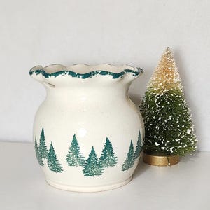 May include: A cream-colored ceramic vase with a scalloped rim and green trim. The vase is decorated with a repeating pattern of green Christmas trees. A small, decorative Christmas tree is next to the vase.