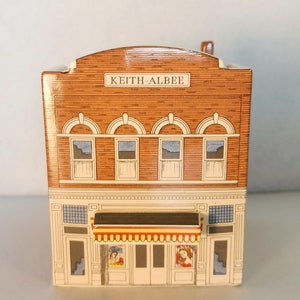 May include: A miniature model of the Keith Albee Theatre, a brick building with a yellow awning and four windows. The model is made of paper and has a detailed design.